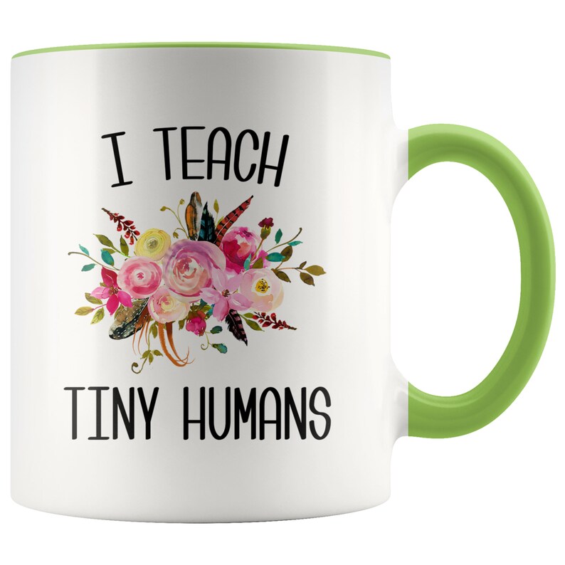 Peut inclure: Une tasse &agrave; caf&eacute; en c&eacute;ramique blanche avec un bord et une poign&eacute;e verts. La tasse pr&eacute;sente un motif floral avec des fleurs roses, jaunes et vertes et des plumes. Le texte "I teach tiny humans" est imprim&eacute; en noir sur la tasse.