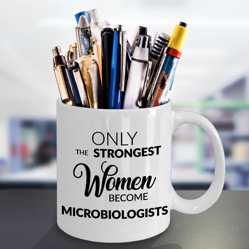 May include: White ceramic mug filled with pens and pencils. The mug has the text "ONLY THE STRONGEST Women BECOME MICROBIOLOGISTS" in black script. The mug is on a gray surface.