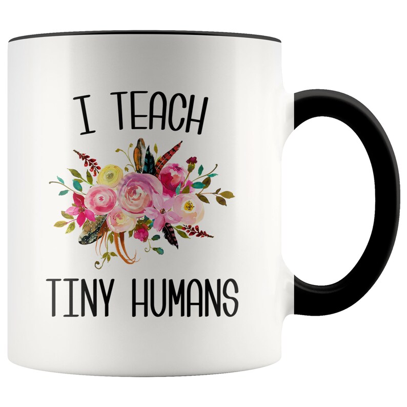 Peut inclure: Une tasse &agrave; caf&eacute; en c&eacute;ramique blanche avec un bord et une poign&eacute;e noirs. La tasse pr&eacute;sente un motif floral avec des fleurs roses, jaunes et vertes et des plumes. Le texte "I teach tiny humans" est imprim&eacute; en noir sur la tasse.