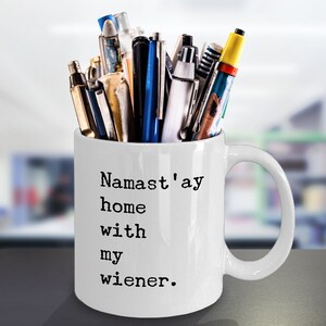 May include: White ceramic mug with a black inscription that reads "Namaste'ay home with my wiener." The mug is filled with a variety of pens and pencils.