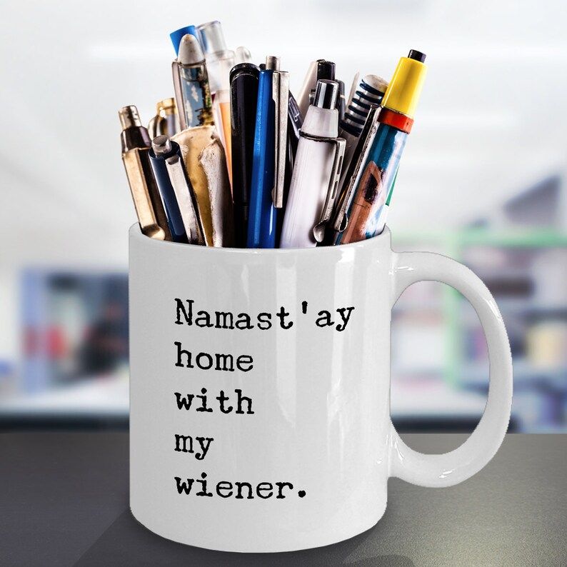 May include: White ceramic mug with a black inscription that reads "Namaste'ay home with my wiener." The mug is filled with a variety of pens and pencils.