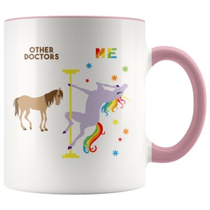 May include: White ceramic coffee mug with a pink rim and handle. The mug features a graphic of a unicorn with a rainbow tail, dancing on a pole. The text "OTHER DOCTORS" is on the left side of the mug and "ME" is on the right side.