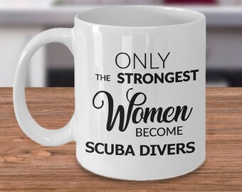 May include: White ceramic mug with the text "ONLY THE STRONGEST Women BECOME SCUBA DIVERS" in black. The mug has a curved handle and is set on a wooden surface.