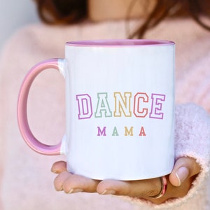May include: White ceramic mug with a pink rim and handle. The mug has the text "DANCE MAMA" in colorful letters.