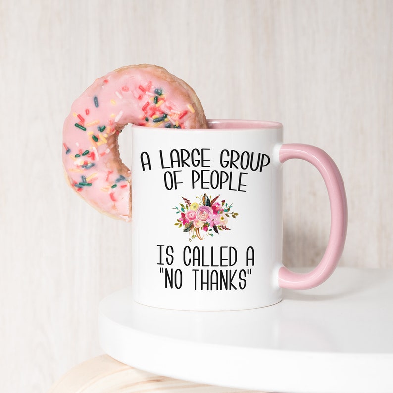 May include: White ceramic mug with a pink handle and the text "A large group of people is called a "no thanks"" printed in black. A pink donut with sprinkles is partially in the mug.