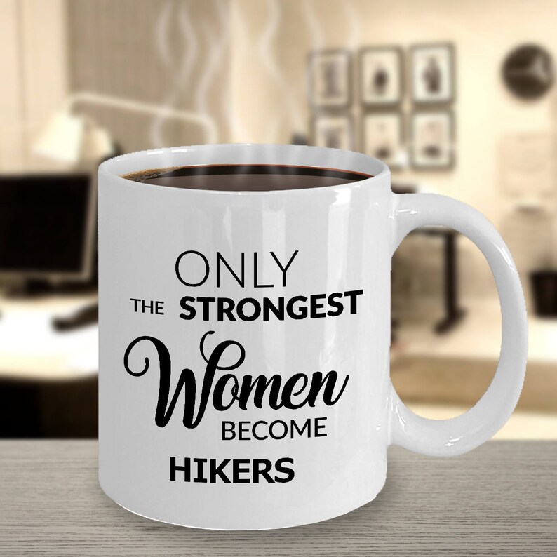 May include: White ceramic mug filled with dark liquid, featuring the text "ONLY THE STRONGEST Women BECOME HIKERS" in black. The mug has a curved handle and is set against a blurred indoor background.