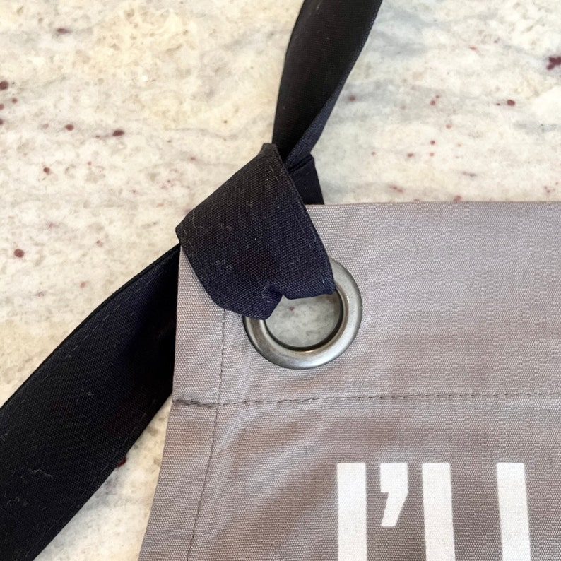 May include: A gray canvas apron with black straps and a metal grommet. The apron has the text "I'll be back" printed on the front.