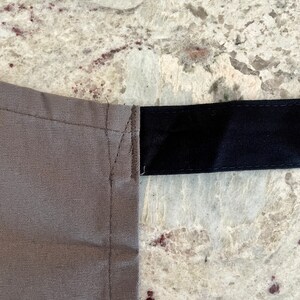 May include: A close-up of a gray canvas fabric with a black strap sewn across the top. The fabric has a subtle texture and the strap is smooth.
