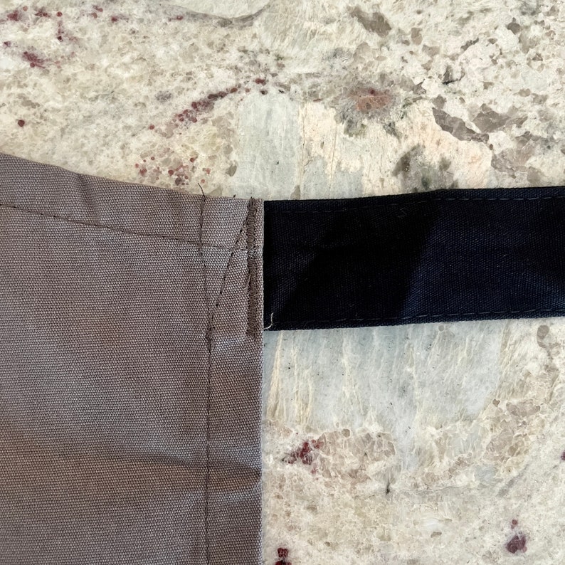 May include: A close-up of a gray canvas fabric with a black strap sewn across the top. The fabric has a subtle texture and the strap is smooth.