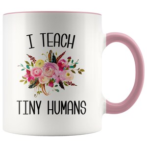 Peut inclure: Une tasse &agrave; caf&eacute; en c&eacute;ramique blanche avec un bord et une poign&eacute;e roses. La tasse pr&eacute;sente un motif floral avec le texte "J'enseigne de petits humains".