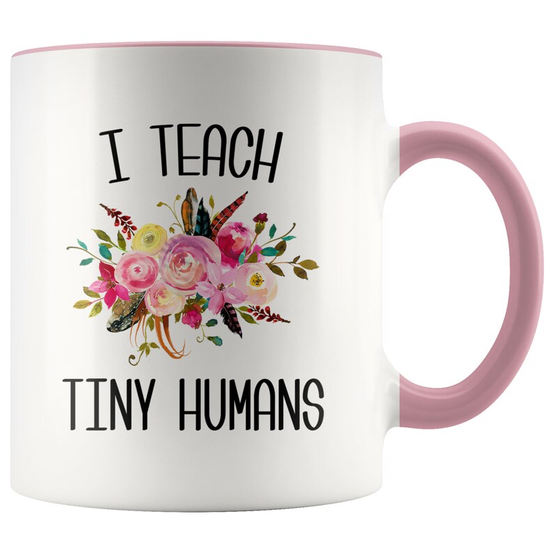 Peut inclure: Une tasse &agrave; caf&eacute; en c&eacute;ramique blanche avec un bord et une poign&eacute;e roses. La tasse pr&eacute;sente un motif floral avec le texte "J'enseigne de petits humains".