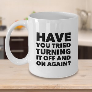 May include: A white ceramic mug with a curved handle. The mug features the black text: "HAVE YOU TRIED TURNING IT OFF AND ON AGAIN?" The mug is sitting on a light-colored wooden surface.