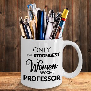 K&ouml;nnte beinhalten: Eine wei&szlig;e Keramik-Kaffeetasse mit schwarzem Text, der "Only the strongest women become professor" lautet. Die Tasse ist mit verschiedenen Stiften und Bleistiften gef&uuml;llt.