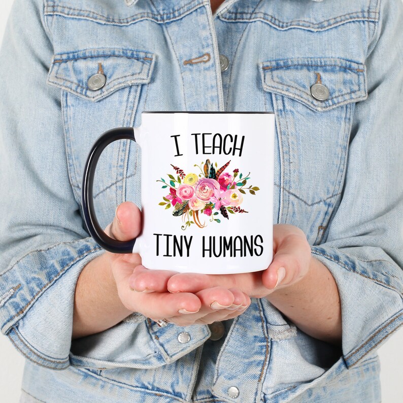 Peut inclure: Une tasse &agrave; caf&eacute; blanche avec une anse noire. La tasse est d&eacute;cor&eacute;e d'un motif floral et du texte "J'enseigne les petits humains".