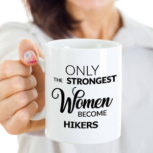 May include: White ceramic mug with black text that reads "ONLY THE STRONGEST Women BECOME HIKERS". The mug is held by a person. The mug is a standard size for coffee or tea.