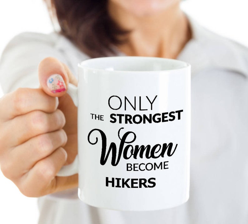 May include: White ceramic mug with black text that reads "ONLY THE STRONGEST Women BECOME HIKERS". The mug is held by a person. The mug is a standard size for coffee or tea.
