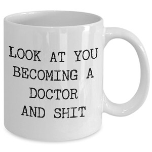 May include: White ceramic mug with the text "LOOK AT YOU BECOMING A DOCTOR AND SHIT" in black, bold, uppercase letters. The mug has a curved handle and a glossy finish.