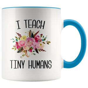 Peut inclure: Une tasse &agrave; caf&eacute; en c&eacute;ramique blanche avec un bord et une poign&eacute;e bleus. La tasse pr&eacute;sente un motif floral et le texte "J'enseigne de petits humains".