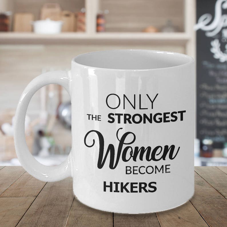 May include: White ceramic mug with a curved handle. The mug features the text "ONLY THE STRONGEST Women BECOME HIKERS" in black lettering. The mug is on a wooden surface.