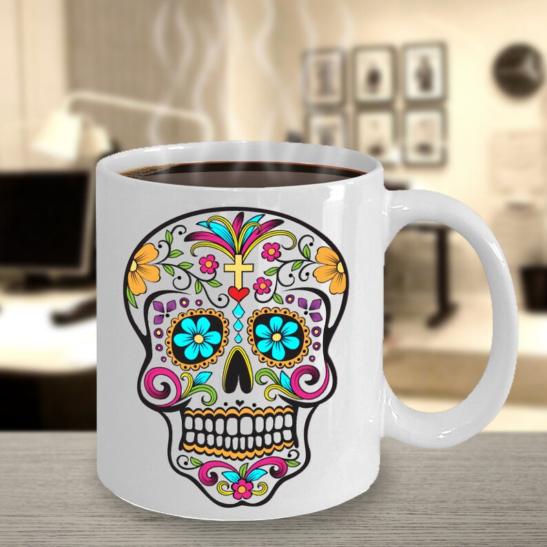 May include: White ceramic coffee mug with a colorful sugar skull design. The skull has blue eyes, yellow teeth, and pink, yellow, and purple flowers.