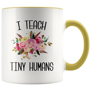 Peut inclure: Une tasse &agrave; caf&eacute; en c&eacute;ramique blanche avec un bord et une poign&eacute;e jaunes. La tasse pr&eacute;sente un motif floral et le texte "J'enseigne de petits humains".