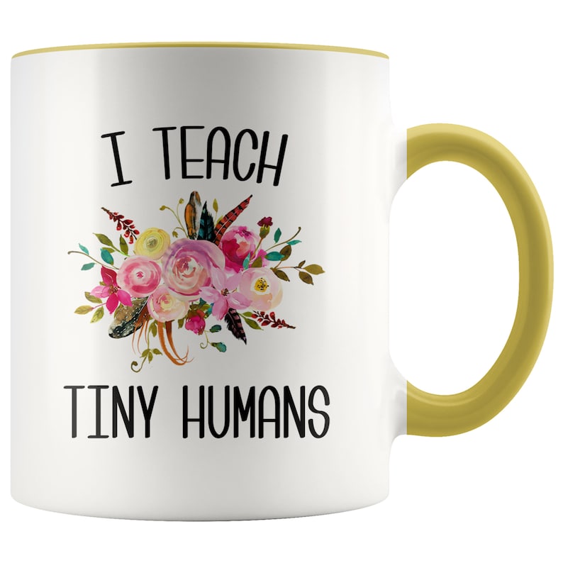 Peut inclure: Une tasse &agrave; caf&eacute; en c&eacute;ramique blanche avec un bord et une poign&eacute;e jaunes. La tasse pr&eacute;sente un motif floral et le texte "J'enseigne de petits humains".