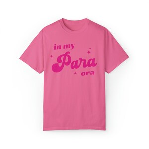 May include: A bright pink t-shirt with the phrase "in my Para era" in a retro font. The text is in a darker shade of pink and is accompanied by star-shaped graphics. The shirt has short sleeves and a crew neck.