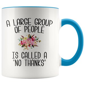 May include: White ceramic coffee mug with a blue rim and handle. The mug features a floral design and the text "A large group of people is called a "no thanks""