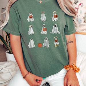 Peut inclure: Un t-shirt vert avec un motif de chiens habill&eacute;s en fant&ocirc;mes tenant des citrouilles. Les chiens sont dans diff&eacute;rentes poses, certains sont assis, d'autres debout et d'autres marchent. Les fant&ocirc;mes sont blancs et les citrouilles sont orange. Le t-shirt est confortable et d&eacute;contract&eacute;.