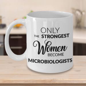 May include: White ceramic mug with a curved handle. The mug features the text "ONLY THE STRONGEST Women BECOME MICROBIOLOGISTS" in black lettering. The mug is sitting on a light brown wooden surface.
