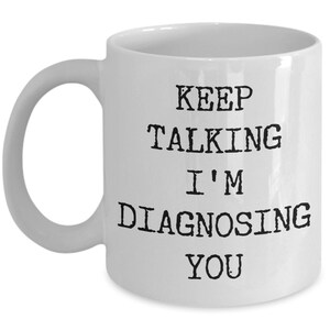 May include: White ceramic coffee mug with black text that reads "Keep Talking I'm Diagnosing You".