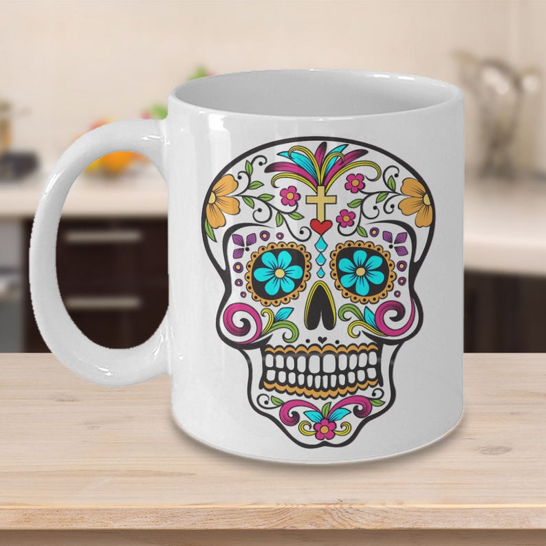 May include: White ceramic coffee mug with a colorful sugar skull design. The skull is decorated with flowers, swirls, and a cross.