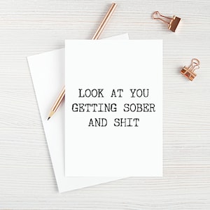 May include: White card with black text that reads "LOOK AT YOU GETTING SOBER AND SHIT". The card is on a white wooden surface with a rose gold pencil and two rose gold paper clips.