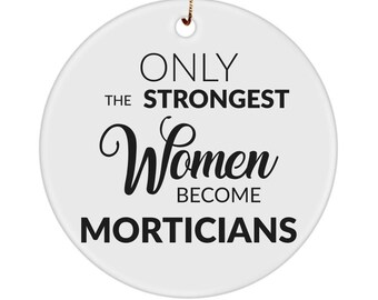 May include: A round white ceramic ornament featuring the black printed text ONLY THE STRONGEST Women BECOME MORTICIANS. The ornament has a small hole at the top for hanging and includes a gold-colored string attached through the opening.