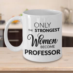 K&ouml;nnte beinhalten: Wei&szlig;e Keramik-Kaffeetasse mit schwarzem Text, der "Only the strongest women become professor" lautet.