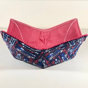 May include: A square bowl cozy with a navy blue exterior patterned with white and red arrows. The interior is red with a white polka dot pattern. The bowl is folded to create a bowl shape.