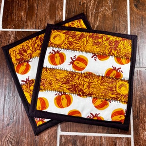 May include: Two square pot holders with a patchwork design. The top and bottom sections feature a yellow and brown floral pattern, while the middle section displays orange pumpkins on a white background. Each pot holder has a black border.