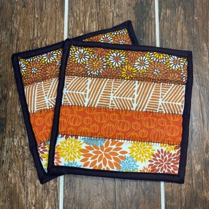 May include: Two square quilted pot holders with a fall theme. The fabric features patterns of pumpkins, leaves, and floral designs in orange, yellow, and white. The edges are trimmed with a dark brown binding.