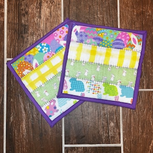 May include: Two purple-trimmed pot holders with an Easter theme. One pot holder features a yellow and white checkered pattern with bunnies and eggs. The other pot holder features a green and white checkered pattern with bunnies and eggs.
