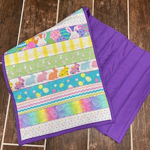 May include: A purple and white table runner with a patchwork design featuring bunnies, eggs, and stripes. The runner is made of fabric and has a quilted texture.