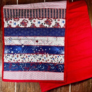May include: A red, white, and blue quilted table runner with a patriotic theme. The runner features a variety of designs, including stars, fireworks, and red trucks. The runner is perfect for adding a touch of Americana to any table.