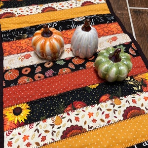 May include: A fall-themed table runner with a patchwork design featuring pumpkins, sunflowers, and autumn leaves in orange, yellow, and black. Three ceramic pumpkin figurines in orange, iridescent white, and green sit on top.