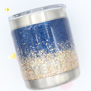 May include: A stainless steel Yeti tumbler with a blue and gold glitter ombre design. The tumbler has a clear lid and the Yeti logo is visible on the side.
