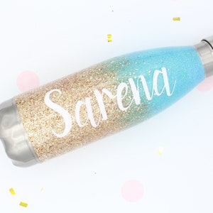 Puede incluir: Una botella de agua personalizada con un dise&ntilde;o degradado de purpurina dorada y azul. El nombre "Sarena" est&aacute; escrito en blanco en la botella.