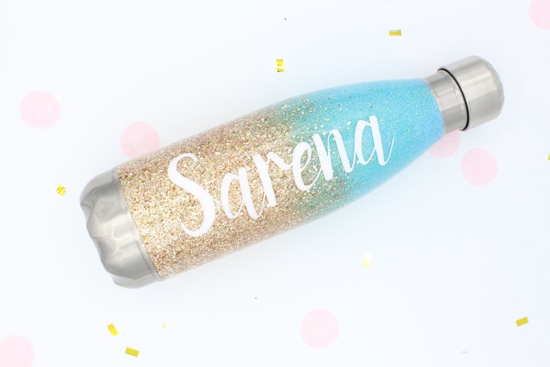 Puede incluir: Una botella de agua personalizada con un dise&ntilde;o degradado de purpurina dorada y azul. El nombre "Sarena" est&aacute; escrito en blanco en la botella.