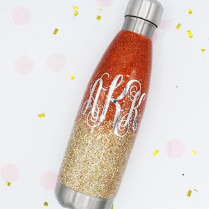 Puede incluir: Una botella de agua de acero inoxidable con un dise&ntilde;o degradado de purpurina dorada y naranja. La botella tiene un monograma con la inicial "J.R.K." en la parte delantera.