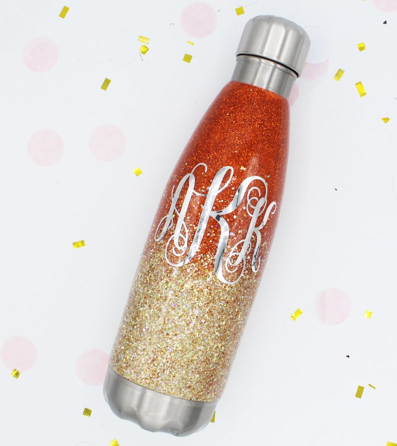 Puede incluir: Una botella de agua de acero inoxidable con un dise&ntilde;o degradado de purpurina dorada y naranja. La botella tiene un monograma con la inicial "J.R.K." en la parte delantera.