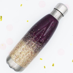 Puede incluir: Una botella de agua de acero inoxidable con un dise&ntilde;o de purpurina degradada en oro y morado. La botella tiene una tapa de rosca.