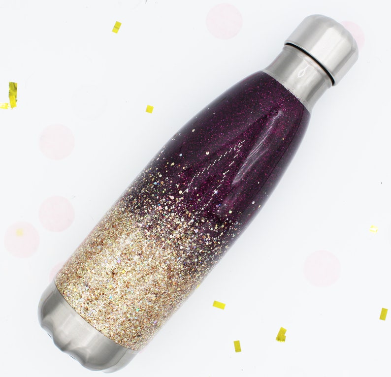 Puede incluir: Una botella de agua de acero inoxidable con un dise&ntilde;o de purpurina degradada en oro y morado. La botella tiene una tapa de rosca.