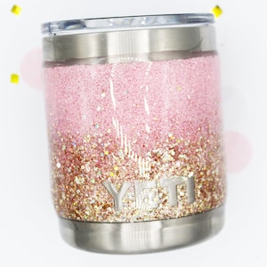 May include: A pink and gold glitter Yeti tumbler with a stainless steel lid and base. The tumbler has a gradient design with a darker pink at the top and a lighter gold at the bottom.